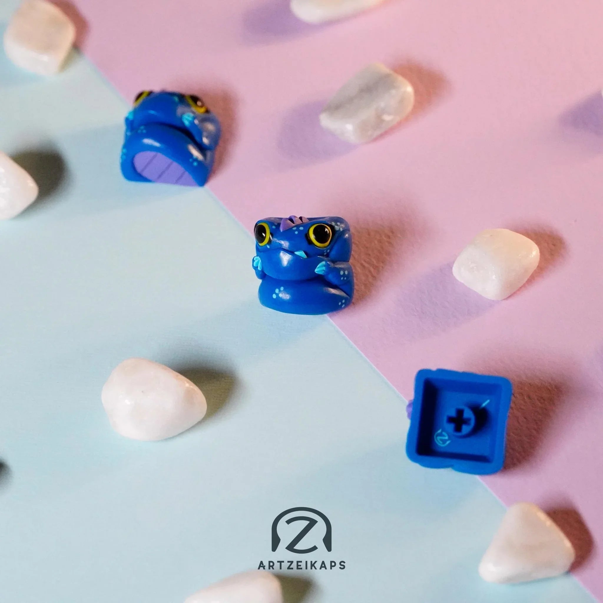 Chomper The Sea Beast artisan keycap trio scattered on pastel blue background with white pebbles, top-down view