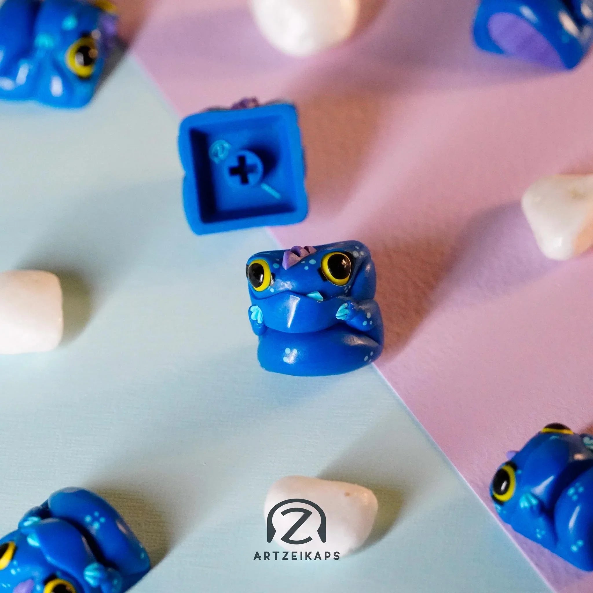 Chomper The Sea Beast artisan keycap in ocean blue with yellow eyes, front and back view on pastel pink and blue background with white pebbles