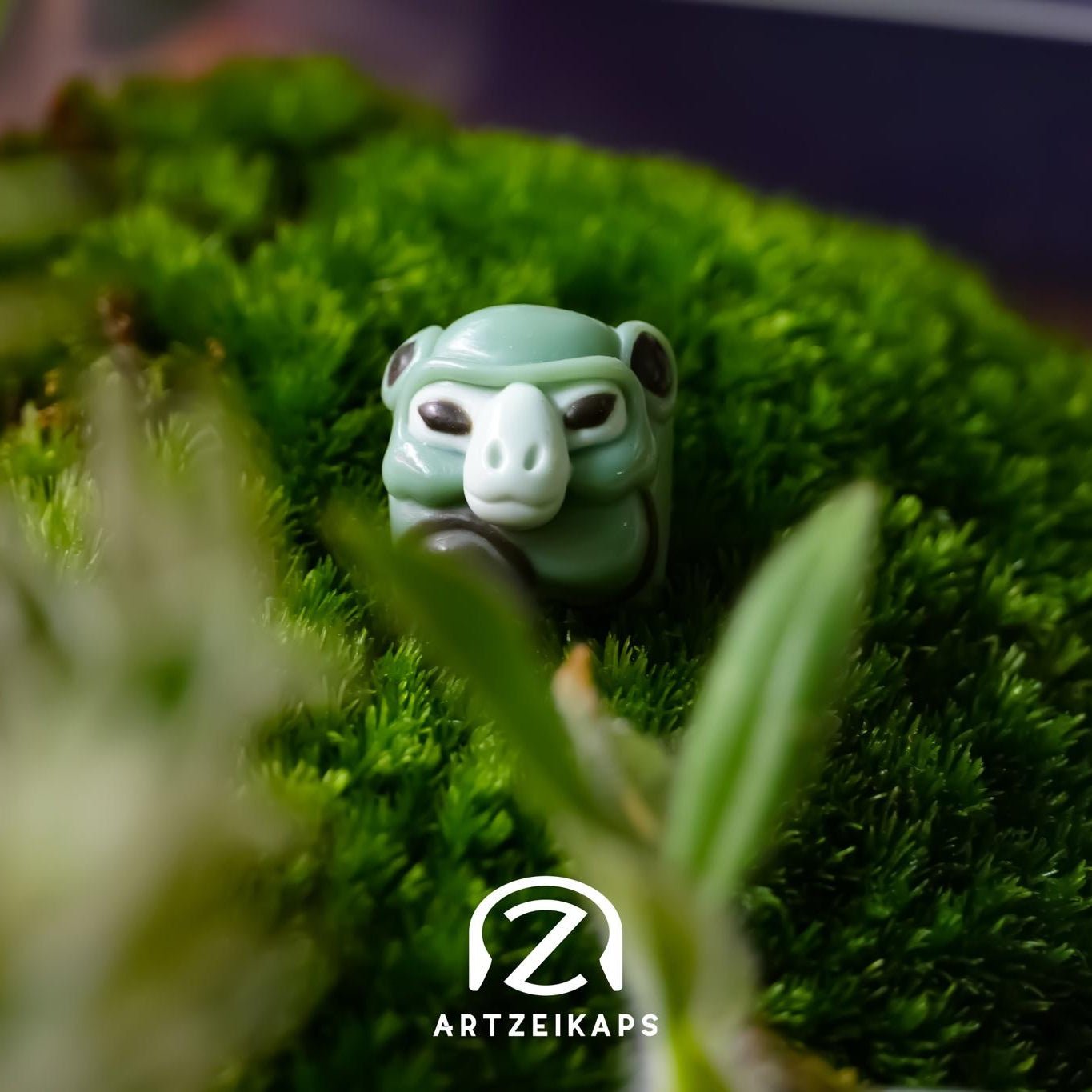 Smunkey GMK November Fog artisan keycap in sage green, solo front view amongst moss and green plants
